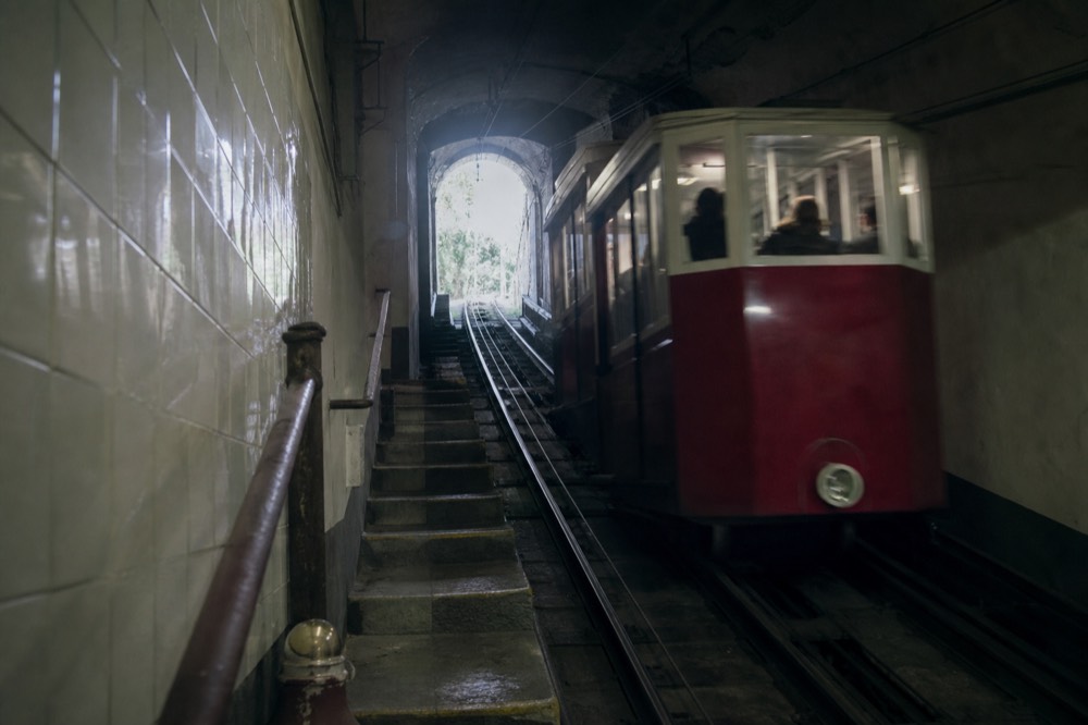 A funicular carriage climbing a steep tunnel track toward daylight, tiled station wall and worn metal handrails in the foreground
