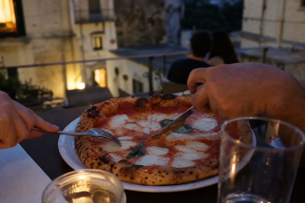 Knife and fork cutting into the soft centre of a margherita pizza on a plain white plate, tomato pooling around the blade