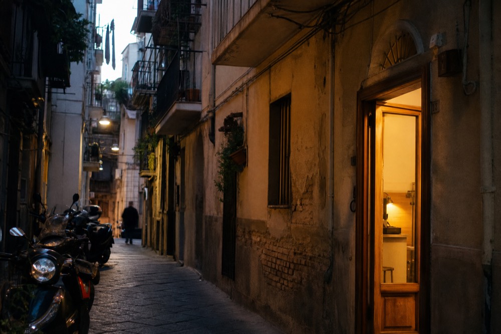 Steep narrow residential alley with balconies, hanging plants, parked scooters and one lit shop doorway glowing at dusk