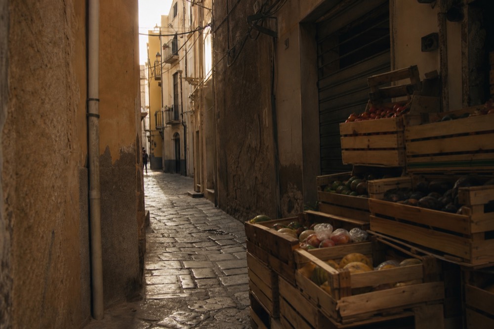 Cracked stone paving and peeling ochre plaster on a residential backstreet, tangled cables overhead, wooden fruit crates stacked in the shade