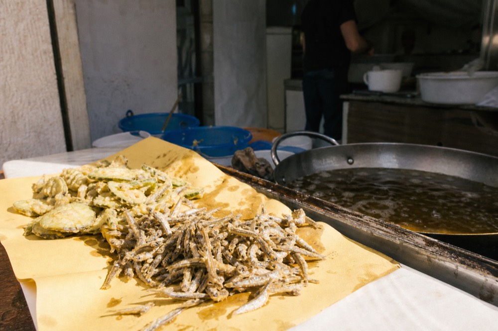 Small fried anchovies and battered vegetables draining on paper beside a deep pan of oil at a tiny street kitchen