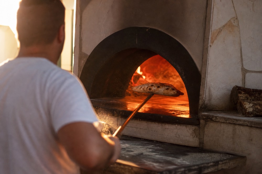 Wood-fired oven mouth glowing orange as a long peel slides a folded pizza out, flour dust visible in the warm light