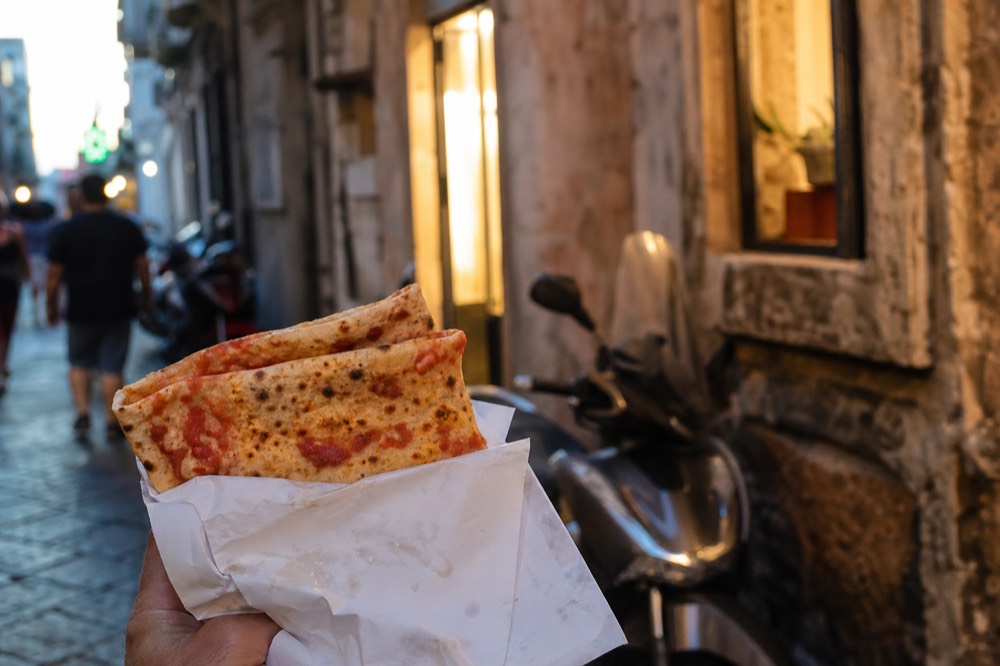 Folded pizza a portafoglio held in one hand on a busy street, grease-spotted paper, blurred scooters behind, no signage visible
