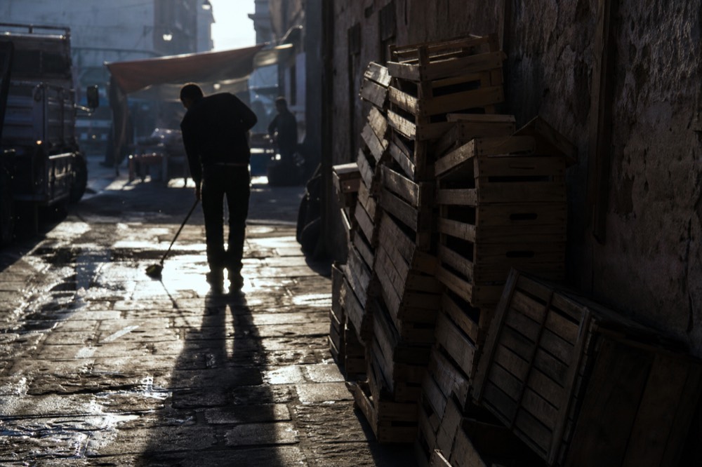 Market street packing up in early afternoon, empty wooden crates stacked against a wall, one vendor sweeping wet stone in low sun