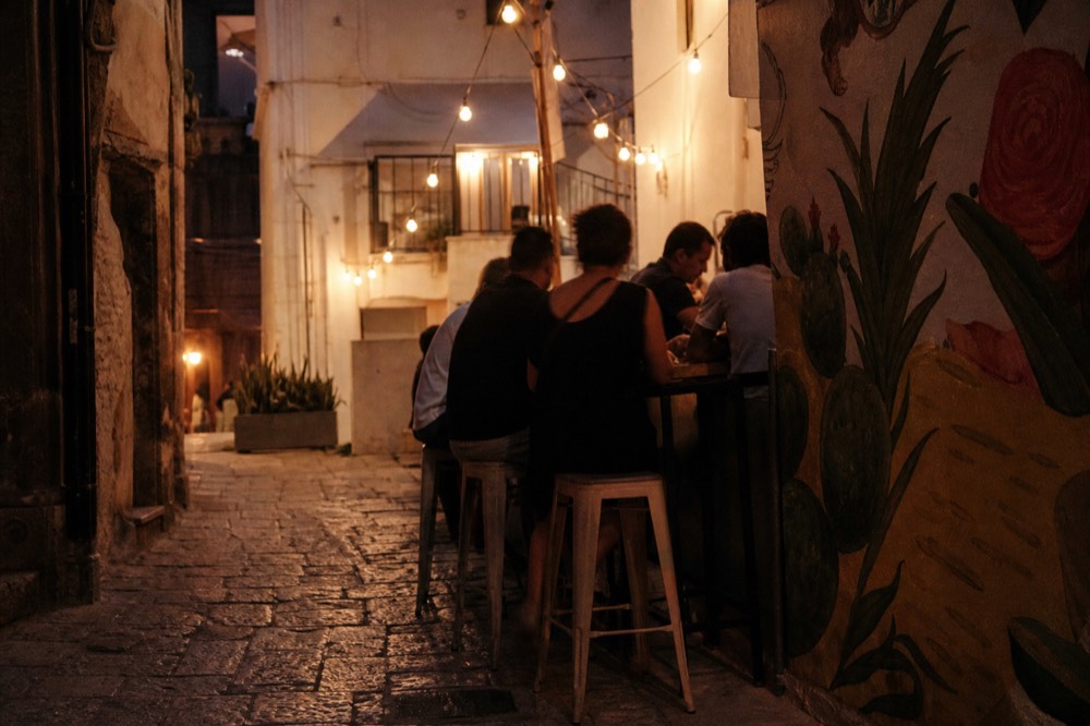 Evening alley with people eating at a small counter under a string of bulbs, worn stone underfoot, mural-painted wall glimpsed at the corner
