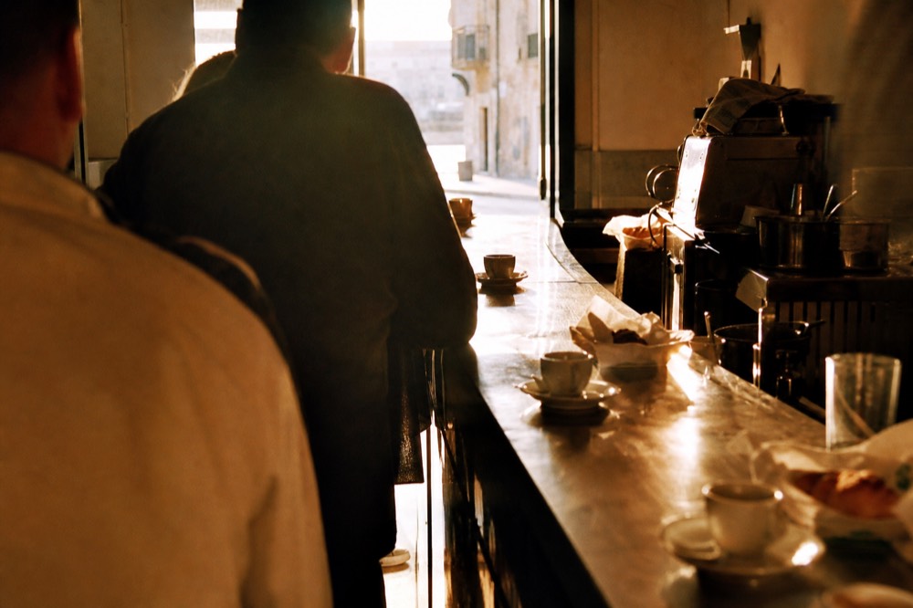 Row of people standing at a zinc bar counter with tiny espresso cups and pastry papers, morning light through an open doorway