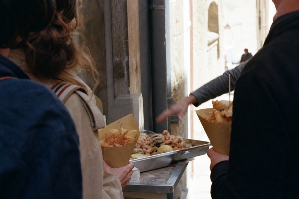 Small group of travelers clustered at a street counter holding paper cones of fried food, guide gesturing toward a tray, sunlit alley behind