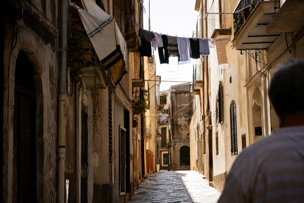 Narrow street canyon of tall shuttered buildings with laundry lines overhead, midday light falling in one bright stripe on the stones