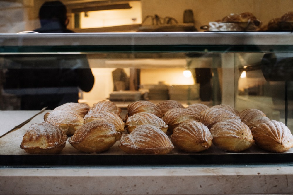 Tray of ridged shell-shaped pastries dusted with icing sugar cooling on baking paper behind a glass counter, warm bakery light