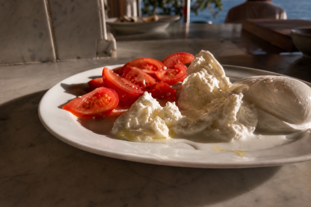 Torn ball of fresh buffalo mozzarella on a white plate weeping whey, sliced tomatoes and a drizzle of olive oil beside it on a marble slab