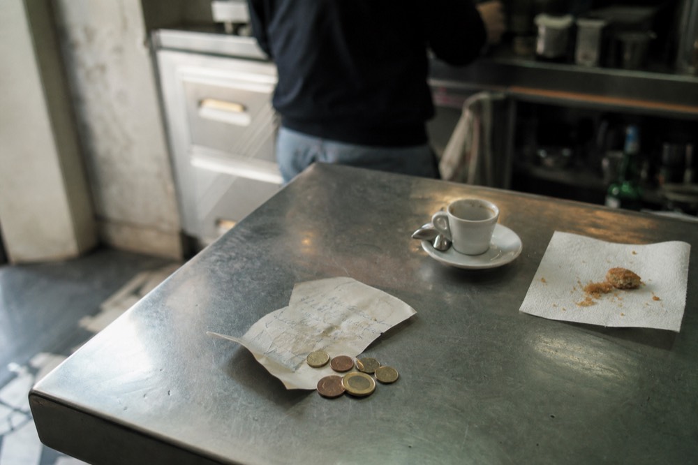 Handwritten paper receipt and coins on a zinc bar counter beside an empty espresso cup and a pastry crumb-strewn napkin